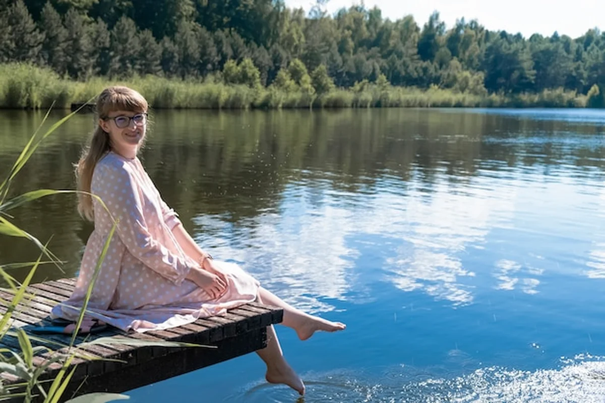 Młoda kobieta siedzi na drewnianym molo nad jeziorem i zanurza stopy w wodzie