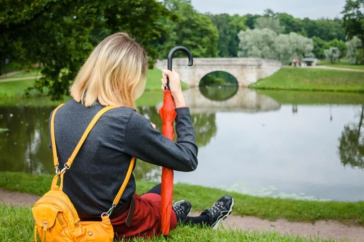 Młoda dziewczyna siedzi na trawie z czerwonym parasolem obok jeziora w parku