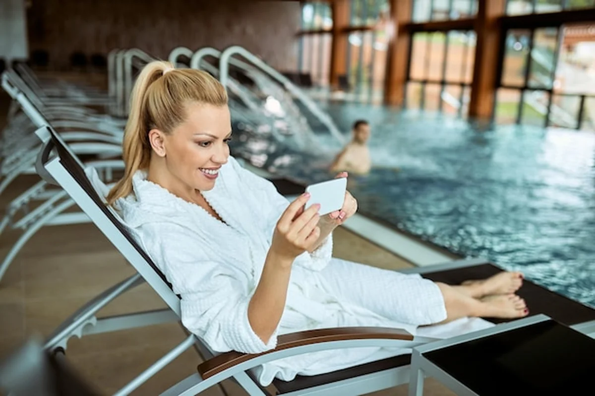 Uśmiechnięta kobieta w szlafroku korzystająca z basenu i telefonu komórkowego w spa